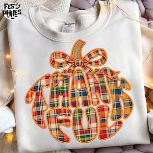 May include: A cream-colored crewneck sweatshirt featuring a large graphic of a pumpkin shape formed by the word THANKFUL in a plaid pattern. The plaid design incorporates red, yellow, green, and blue colors with an orange outline.