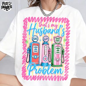 May include: White t-shirt with a colorful graphic featuring three vintage gas pumps in red, pink, and green. The text "that's my Husband's Problem" is written in blue and pink, framed by a pink wavy border.