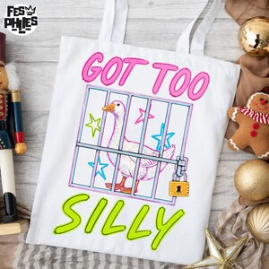 May include: White tote bag with a cartoon goose in a cage, the text "GOT TOO SILLY" in pink and green. The goose is white with an orange beak and feet. The cage has blue and pink stars.