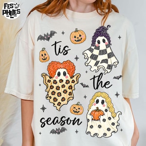 Puede incluir: Una camiseta de color crema con un estampado gráfico que incluye el texto 'tis the season'. El diseño presenta tres fantasmas con diferentes patrones, calabazas naranjas y murciélagos negros dispersos por la parte frontal de la prenda.