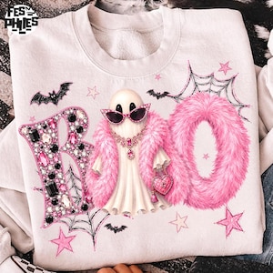 May include: A white sweatshirt featuring a graphic of a ghost wearing pink cat-eye sunglasses, a pink faux fur stole, and a heart-shaped purse. The word BOO is printed with the B in jewels and the OO in pink faux fur, surrounded by bats and stars.