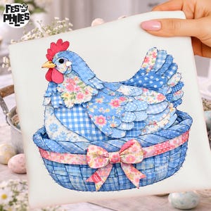 Könnte beinhalten: Eine blau-weiße Patchwork-Henne sitzt in einem blauen Korb mit einer rosa Schleife. Die Henne hat einen roten Kamm und Schnabel und besteht aus verschiedenen gemusterten Stoffen, darunter Karos und Blumendrucke. Der Korb hat ein rosa-weißes Tupfenband.