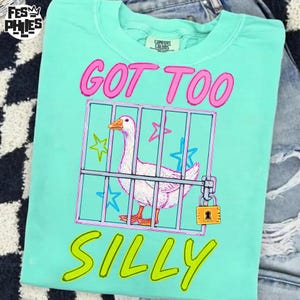 May include: A light teal Comfort Colors t-shirt with a graphic of a white goose inside a pink and blue cage. The text "GOT TOO SILLY" is in pink and yellow. The shirt is folded on top of a pair of jeans.