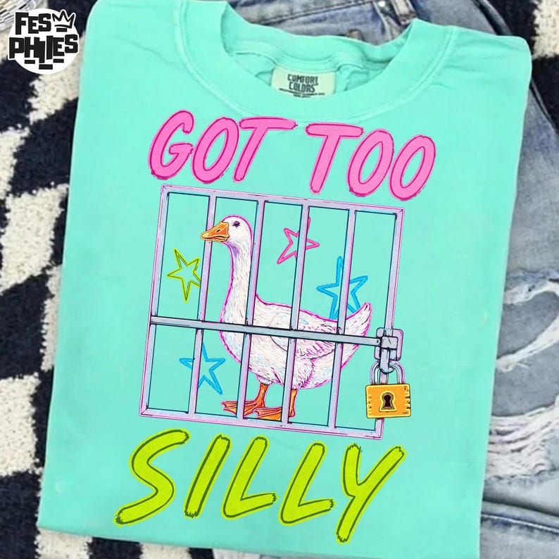 May include: A light teal Comfort Colors t-shirt with a graphic of a white goose inside a pink and blue cage. The text "GOT TOO SILLY" is in pink and yellow. The shirt is folded on top of a pair of jeans.