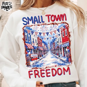 Könnte beinhalten: Weißes Sweatshirt mit patriotischem Design. Es zeigt eine Kleinstadt-Straßenszene in Rot, Weiß und Blau mit den Worten "SMALL TOWN FREEDOM". Das Design umfasst Feuerwerk und amerikanische Flaggen.
