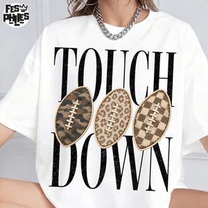May include: A white short-sleeve t-shirt featuring the black text TOUCH DOWN and three football graphics. The footballs display camouflage, leopard print, and checkerboard patterns. A silver chain necklace is worn around the neck area.