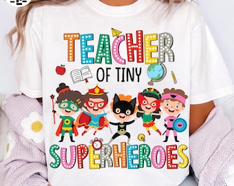May include: White t-shirt with the text "TEACHER OF TINY SUPERHEROES" in colorful letters. The shirt features cartoon illustrations of children dressed as superheroes, along with school-themed graphics like an apple, book, and globe.