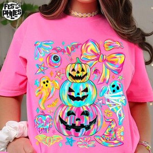 May include: Pink t-shirt featuring a colorful Halloween-themed design. The graphic includes stacked jack-o'-lanterns, ghosts, bats, and a bow, all in vibrant rainbow hues. The shirt has a crew neck and short sleeves.