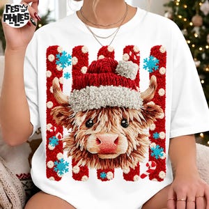 May include: A white t-shirt featuring a graphic of a Highland cow wearing a red and white Santa hat. The cow is centered against a background of vertical red textured stripes with white polka dots, blue snowflakes, and red and white candy canes.
