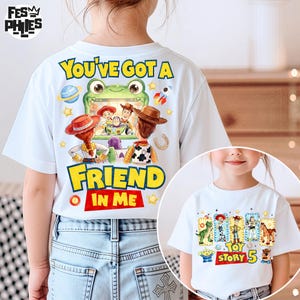 May include: A white short-sleeved t-shirt featuring a graphic of a green frog holding a screen displaying Toy Story characters Woody, Jessie, and Buzz Lightyear with the text You've Got A Friend In Me and a separate Toy Story 5 graphic.