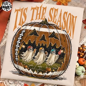 May include: A white t-shirt featuring a large orange pumpkin carved like a football, with the text 'TIS THE SEASON' in orange above. Inside the pumpkin, three ghosts wearing football helmets play on a field under stadium lights.