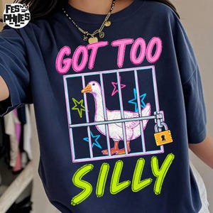 May include: Navy blue t-shirt with a graphic of a white goose inside a cage, with the words "GOT TOO SILLY" in pink and green. The goose has an orange beak and feet. The cage is locked with a gold padlock.