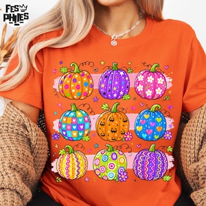 Op de afbeelding: Een oranje T-shirt met korte mouwen en een grafische print van negen kleurrijke pompoenen in een raster. De pompoenen hebben diverse patronen zoals stippen, sterren, bloemen, hartjes en krullen in de kleuren paars, blauw, roze en geel.
