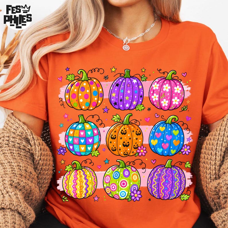 Op de afbeelding: Een oranje T-shirt met korte mouwen en een grafische print van negen kleurrijke pompoenen in een raster. De pompoenen hebben diverse patronen zoals stippen, sterren, bloemen, hartjes en krullen in de kleuren paars, blauw, roze en geel.