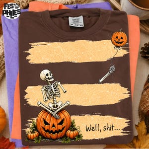 May include: A brown t-shirt featuring a graphic of a skeleton sitting on a large jack-o'-lantern, holding a string attached to a floating jack-o'-lantern balloon. The design includes orange brushstroke accents and the text "Well, shit..."