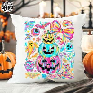May include: White decorative pillow with a colorful Halloween-themed design. The design features three stacked jack-o'-lanterns, ghosts, bats, stars, and a large bow, all in vibrant rainbow hues. The pillow is surrounded by pumpkins.