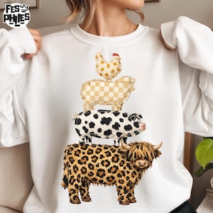 Może przedstawiać: Biała bluza z nadrukiem przedstawiającym ułożone w stos zwierzęta gospodarskie: kurę, owcę, świnię i krowę. Krowa ma wzór w panterkę. Pozostałe zwierzęta mają różne wzory. Bluza ma napis "FES PHIES" w lewym górnym rogu.