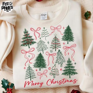 May include: Cream-coloured sweatshirt with a festive design featuring green Christmas trees and red bows. The words "Merry Christmas" are printed in red script at the bottom. A cosy holiday-themed garment.