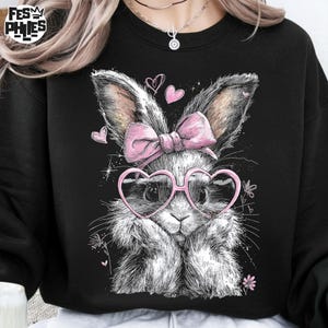 Puede incluir: Sudadera negra con una ilustraci&oacute;n detallada de un conejito con gafas rosas en forma de coraz&oacute;n y un lazo rosa. El conejito tiene pelaje esponjoso y est&aacute; rodeado de peque&ntilde;os corazones y flores. La sudadera tiene el texto "FESM PHIES".