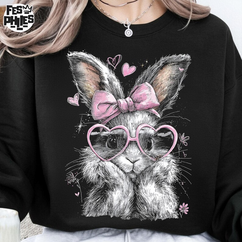 Puede incluir: Sudadera negra con una ilustraci&oacute;n detallada de un conejito con gafas rosas en forma de coraz&oacute;n y un lazo rosa. El conejito tiene pelaje esponjoso y est&aacute; rodeado de peque&ntilde;os corazones y flores. La sudadera tiene el texto "FESM PHIES".