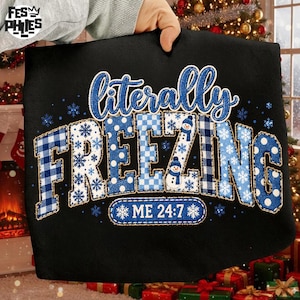 May include: A black sweatshirt featuring the text literally FREEZING ME 24:7 in blue and white patterns. The letters include plaid, polka dot, and snowflake designs, with small snowman graphics integrated into the word FREEZING.