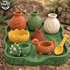 Puede incluir: Un juego de utensilios de cocina de silicona con temática de dinosaurios que incluye tres jarrones pequeños, un cuenco de huevo roto, un molde de volcán, un cuenco de triceratops, un cuenco de dinosaurio y una cuchara sobre una bandeja verde.