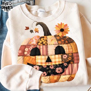 May include: Cream-colored sweatshirt featuring a patchwork pumpkin design. The pumpkin has a smiling face and is decorated with floral and plaid patterns in shades of orange, brown, and pink. A sunflower and leaves are also included.