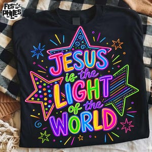 May include: A black t-shirt featuring a vibrant, multi-colored graphic design with the text "JESUS is the LIGHT of the WORLD." The design includes three large stars in neon pink, blue, and green, surrounded by smaller star and line accents.