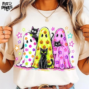 May include: A cream-colored t-shirt featuring a graphic of three ghosts and two black cats. One ghost has colorful polka dots, one is lime green with orange pumpkins, and one is pink with flowers. Brightly colored stars surround the figures.