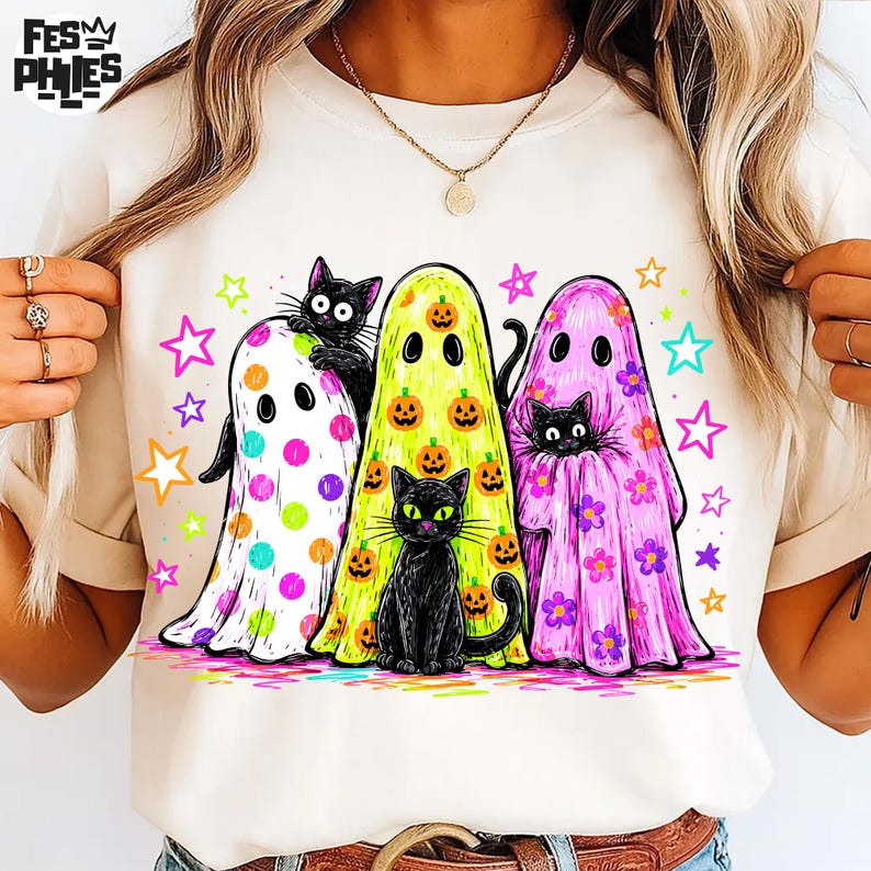 May include: A cream-colored t-shirt featuring a graphic of three ghosts and two black cats. One ghost has colorful polka dots, one is lime green with orange pumpkins, and one is pink with flowers. Brightly colored stars surround the figures.