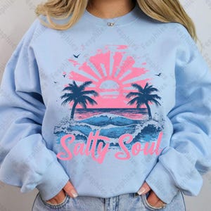 May include: A light blue sweatshirt featuring a graphic design of a sunset over the ocean with palm trees and the text "Salty Soul" in pink.