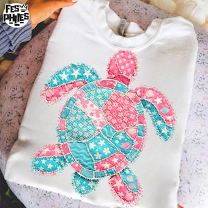 Puede incluir: Sudadera blanca con un diseño de tortuga marina de patchwork. La tortuga está hecha de tela rosa y turquesa con patrones de estrellas y flores. El diseño está centrado en la parte delantera de la sudadera.