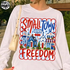 Könnte beinhalten: Weißes Langarmshirt mit einem farbenfrohen Grafikdesign. Das Design enthält die Worte "SMALL TOWN FREEDOM" und Illustrationen von Gebäuden. Das Design verwendet rote, weiße und blaue Farben.