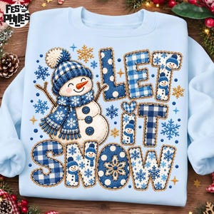 May include: A light blue crewneck sweatshirt featuring a graphic of a snowman wearing a blue and white plaid hat and scarf. The text "LET IT SNOW" is displayed in large, blue and white plaid and polka dot letters, accented with small snowflakes.