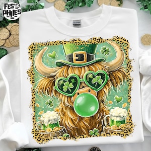 May include: White sweatshirt featuring a cartoon Highland cow wearing heart-shaped shamrock sunglasses and a green leprechaun hat. The cow is blowing a green bubble and surrounded by beer mugs and gold coins. The design has a leopard print border.