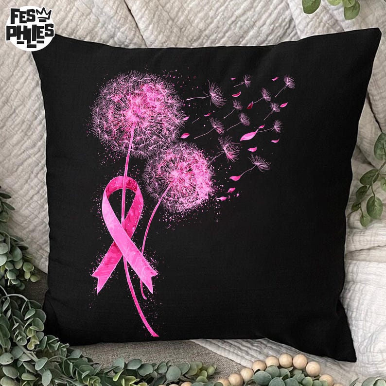 May include: Black square pillow featuring a pink ribbon and two pink dandelions with seeds blowing in the wind. The pillow is a decorative item, likely for a bed or couch.