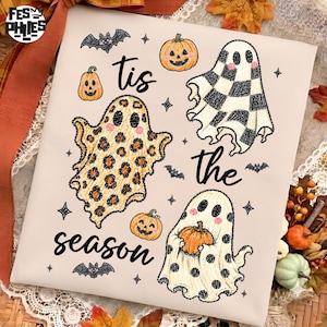 May include: A light beige t-shirt featuring a Halloween graphic with the text 'tis the season'. The design includes three ghosts with different patterns: leopard print, black and white checkerboard, and polka dots, alongside small orange pumpkins and bats.