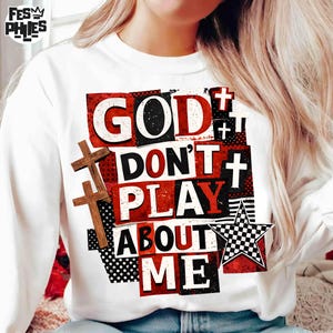 May include: White sweatshirt with the text "GOD DON'T PLAY ABOUT ME" in a bold, layered design. The design includes red and black blocks, wooden crosses, and a checkered star, creating a striking visual statement.