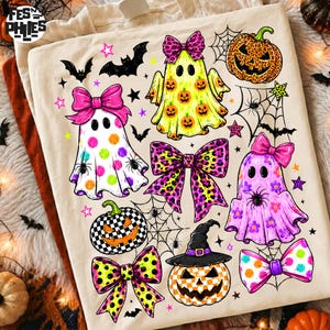 May include: A cream-colored t-shirt featuring a Halloween-themed graphic print. The design includes three ghosts with bows, two jack-o'-lanterns, spiderwebs, bats, and stars in vibrant pink, yellow, orange, and green colors with leopard patterns.