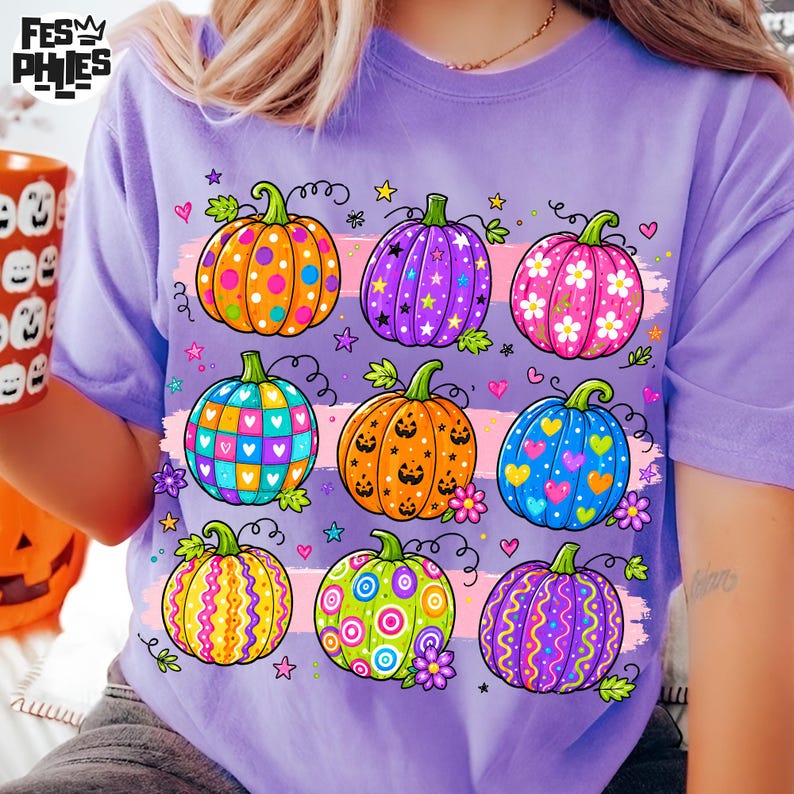 Op de afbeelding: Een lichtpaars t-shirt met een grafische print van negen kleurrijke pompoenen met diverse patronen zoals stippen, sterren, bloemen, hartjes en krullen, geaccentueerd door kleine sterretjes en hartjes, met de tekst FES PHIES in de hoek.