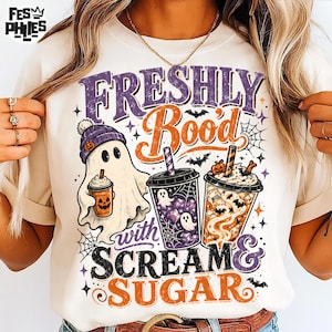 May include: A cream-colored t-shirt featuring a graphic with the text "FRESHLY Boo'd with SCREAM & SUGAR". The design includes a ghost wearing a purple beanie holding a pumpkin cup, alongside two iced coffee cups with ghost and bat illustrations.