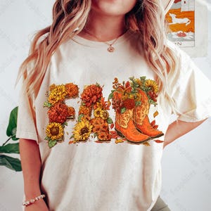 May include: Cream-coloured t-shirt featuring the word "FALL" in a floral design. The letters are filled with sunflowers, orange and yellow mums, and autumn leaves. Two orange cowboy boots are filled with flowers.