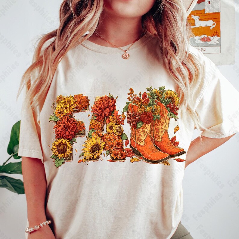 May include: Cream-coloured t-shirt featuring the word "FALL" in a floral design. The letters are filled with sunflowers, orange and yellow mums, and autumn leaves. Two orange cowboy boots are filled with flowers.