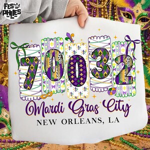 May include: A white cushion featuring the text "70032 Mardi Gras City New Orleans, LA". The numbers are decorated with purple, green, and gold patterns and fleur-de-lis symbols. The cushion has a scalloped edge and is themed for Mardi Gras.