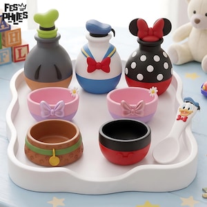 May include: A set of Disney-themed kitchenware on a white tray, including three character-shaped bottles, two pink bowls with bows, a brown bowl with a paw print pattern, a black and red bowl, and a white spoon with a Donald Duck character head.