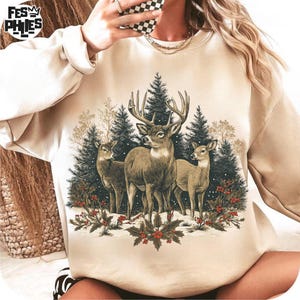 May include: Beige sweatshirt featuring a detailed illustration of three deer in a winter forest scene. The design includes evergreen trees, holly with red berries, and a snowy ground. The sweatshirt has a crew neck.