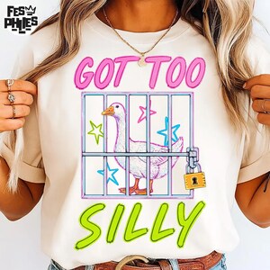 May include: Cream-colored t-shirt with the text "GOT TOO SILLY" in pink and green. A cartoon goose is inside a cage with stars. The cage has a padlock.