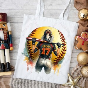 May include: White tote bag with a watercolor design of a softball player with the name "Emma" and the number "17" on the back of their jersey.