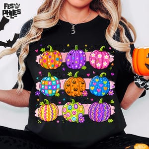 Op de afbeelding: Een zwart t-shirt met een grafische print van negen pompoenen gerangschikt in drie rijen. De pompoenen zijn versierd met kleurrijke patronen, waaronder stippen, sterren, bloemen, hartjes en krullen in de kleuren oranje, paars, roze, blauw en groen.