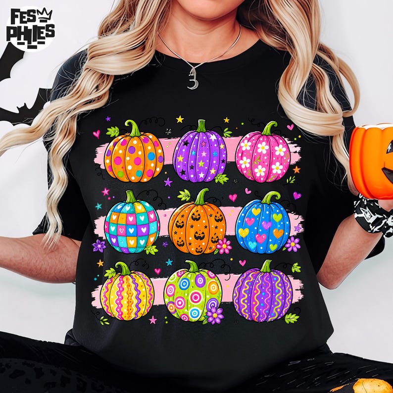 Op de afbeelding: Een zwart t-shirt met een grafische print van negen pompoenen gerangschikt in drie rijen. De pompoenen zijn versierd met kleurrijke patronen, waaronder stippen, sterren, bloemen, hartjes en krullen in de kleuren oranje, paars, roze, blauw en groen.
